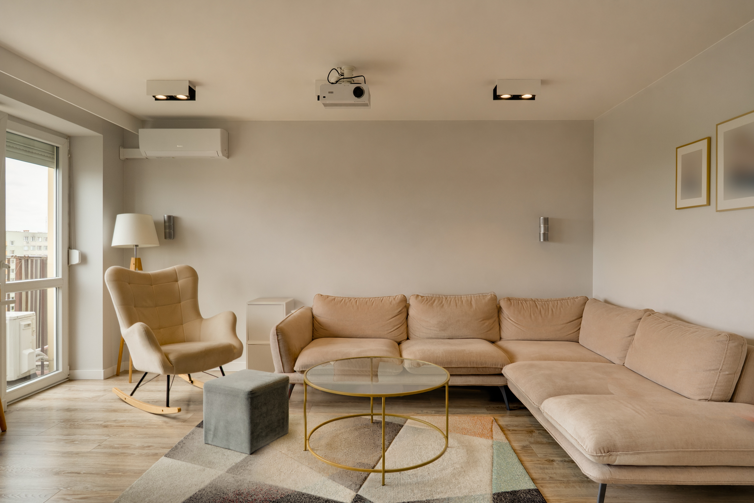 Stylowy salon z beżową sofą i fotelem, z dużym oknem i projektorem sufitowym.